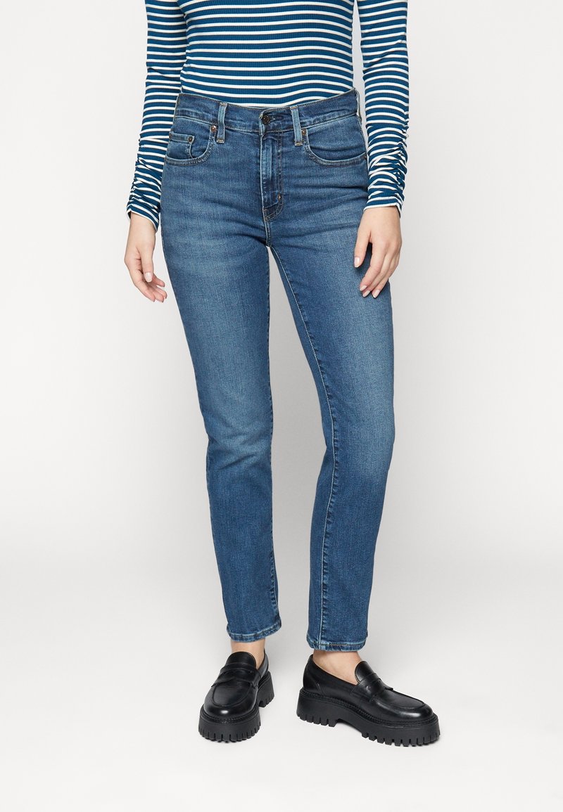 Personne portant un jean bleu taille haute, des mocassins noirs épais et un haut à manches longues rayé bleu et blanc, debout devant un fond blanc.