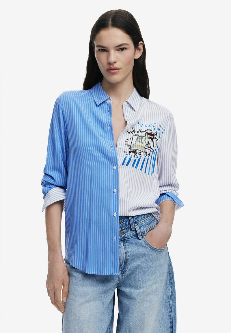 Camicia a righe con un lato blu e un lato bianco, caratterizzata da un design grafico. Tessuto leggero con maniche arrotolate e una vestibilità comoda.