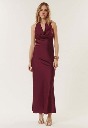 Femme debout portant une robe longue sans manches en satin bordeaux avec un décolleté drapé et des sandales dorées, fond clair uni.