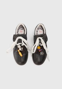 Fekete bőr tornacipő, fehér fűzőkkel, párnázott nyelvvel és különféle díszítő charmokkal a fűzőkön, mintás bélésbéleléssel.