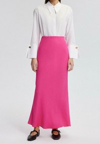 Weißes Hemd mit Kragen, langen Ärmeln und geknöpften Manschetten, kombiniert mit einem figurbetonten pinken Maxirock mit strukturierter Oberfläche. Schwarze Ballerinas.