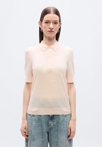 Ανοιχτό ροζ πόλο μπλουζάκι με κοντά μανίκια, γιακά και πέντε κουμπιά, κατασκευασμένο από απαλό, ελαφρύ ύφασμα. Φορεμένο με χαλαρά μπλε τζιν.