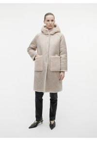 Cappotto in shearling beige con cappucci, dotato di grandi tasche frontali e una texture morbida. Abbinato a pantaloni neri slim-fit e scarpe a punta.