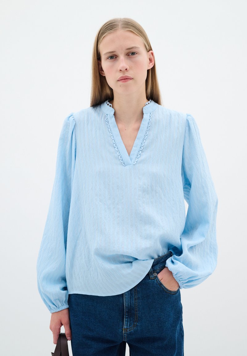 Ljusblå blus med v-ringning, strukturerad tyg och volangdetaljer längs kragen. Har puffärmar och en avslappnad passform.