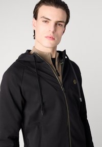 Čierna mikina na zips s textúrovaným povrchom, so svetlo béžovou vnútornou vrstvou, kontrastným žltým prešívaním a logom na hrudi.