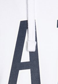 Sweat à capuche blanc arborant une grande lettre graphique "A" bleu marine, avec un tissu côtel é texturé et un cordon blanc.