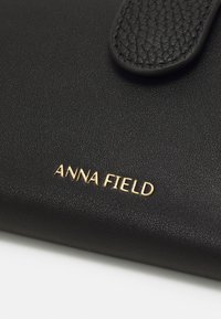 Portofel negru din piele, având o suprafață texturată, închidere cu clapetă și un logo auriu "ANNA FIELD" embosat pe față.