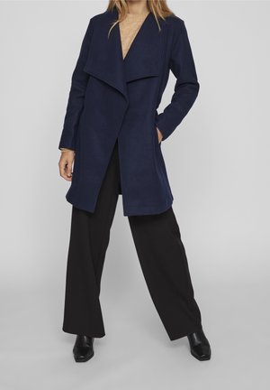 Femme portant un long manteau bleu marine, un haut beige, un pantalon large noir et des bottines noires à bout carré, debout devant un fond clair.