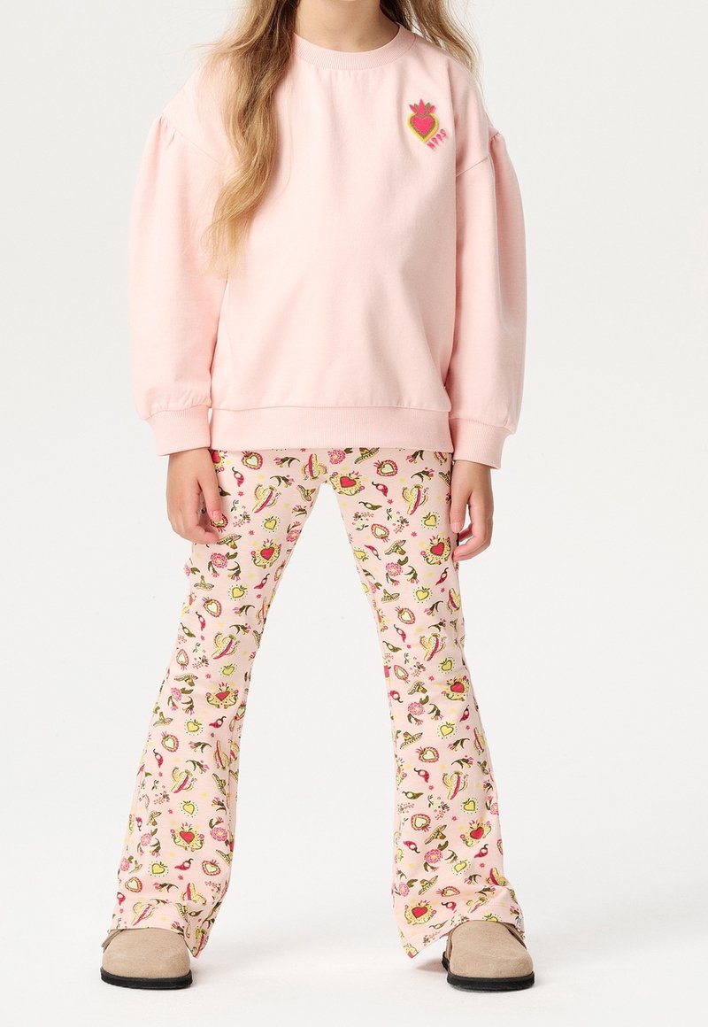 Enfant portant un sweat-shirt rose avec un écusson en forme de cœur et un pantalon évasé assorti à motifs de cœurs et de fleurs, debout les mains détendues.