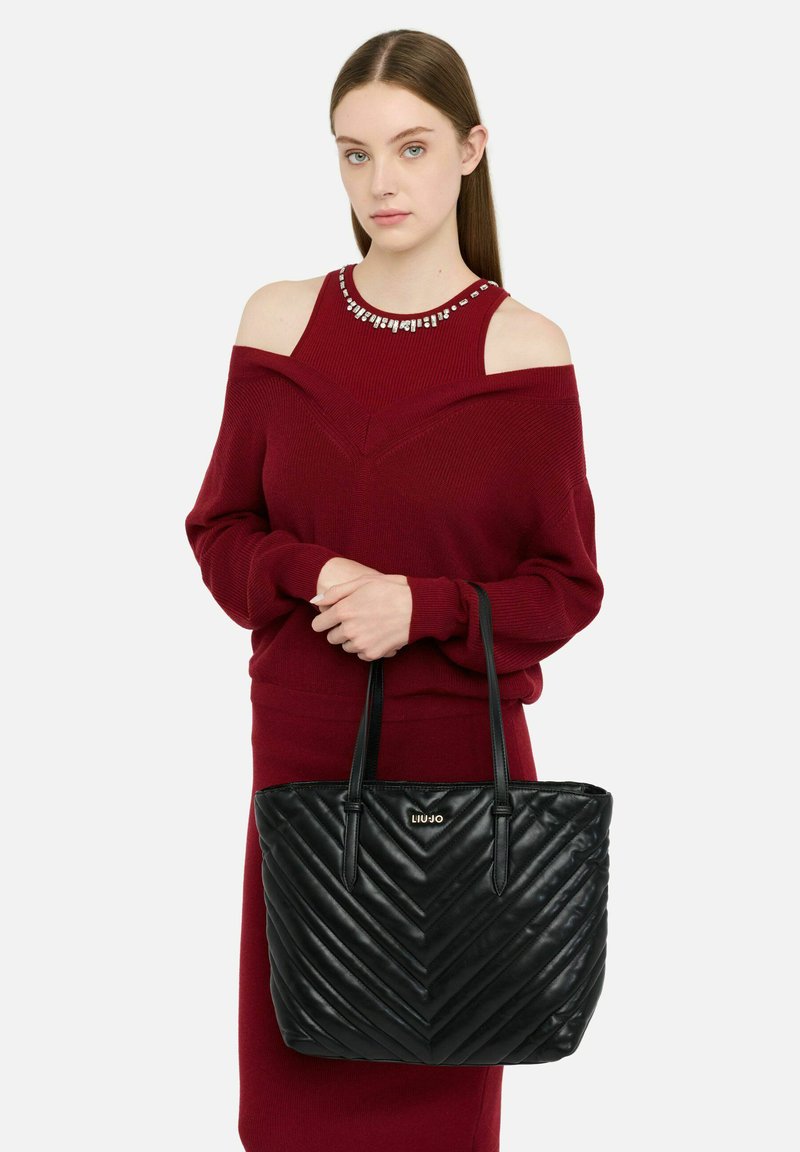 Schwarze gesteppte Tragetasche mit Chevron-Muster und zwei Schulterriemen. Kombiniert mit einem burgunderroten Off-Shoulder-Pullover.