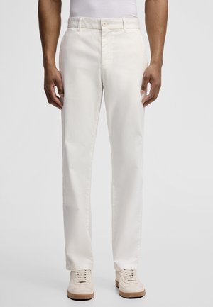 Mann trägt gerade geschnittene, cremefarbene Hosen mit vorderer Knopfleiste und Seitentaschen, kombiniert mit beigen Sneakers und einem weißen Oberteil.