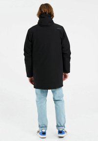 Giacca nera impermeabile con cappuccio, taglio lungo e texture liscia. Indossata sopra jeans azzurri chiari e calzature con accenti di blu e grigio.