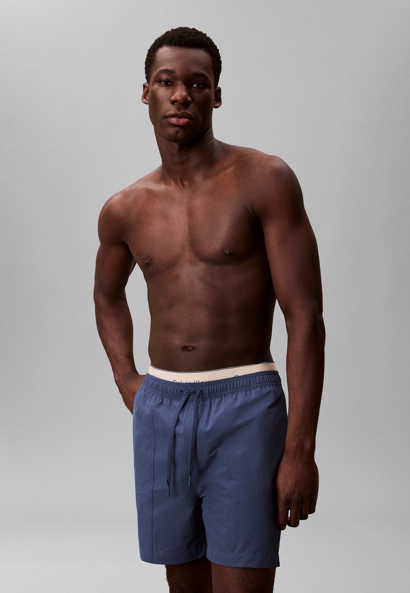 Boxer da bagno blu con vita elasticizzata e lacci, caratterizzati da una texture liscia e un design minimale. Il modello è in piedi con le mani sui fianchi.