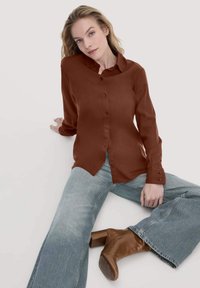 Blusa marrone a maniche lunghe con bottoni frontali, abbinata a jeans svasati blu chiaro e stivali con tacco marroni. Texture liscia, design del colletto classico.