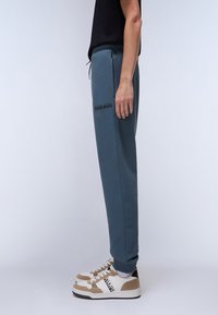 Blue sweatpants with a drawstring waist and side pocket, featuring a black logo detail. Paired with beige and white stylish athletic shoes.