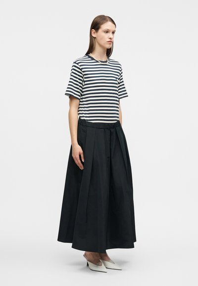 Γυναίκα που φοράει μαύρο παντελόνι με φαρδιά μπατζάκια, άσπρο και μπλε πουά κοντομάνικο πουκάμισο, μαργαριταρένιο κολιέ και λευκές μυτερές γόβες.