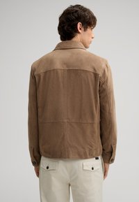 Braune Wildlederjacke mit glatter Textur, präsentiert mit einem einfarbigen Rücken und einem Kragen. Kombiniert mit hellen Hosen und Knopfdetails.