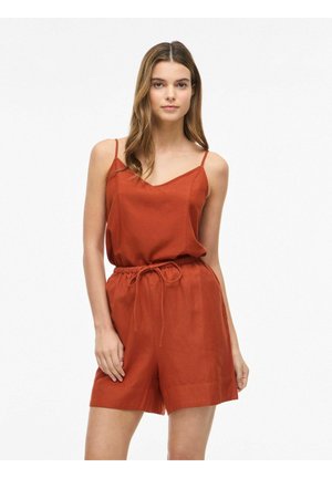 Nő ujjatlan rozsdaszínű-narancssárga overallban, húzózsinóros derékkal, egyszínű fehér háttér előtt áll.