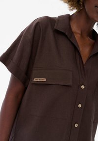 Ciemnobrązowa koszula z krótkim rękawem, zapinana na guziki, z kieszonką na piersi i napisem "blue shadow", noszona przez osobę z kręconymi włosami.