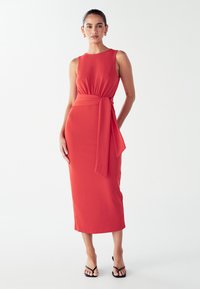 Rotes ärmelloses Kleid mit tailliertem Oberteil, gerafftem Bund und gebundenem Gürtel. Midi-Länge, glatte Textur, kombiniert mit schwarzen Riemchensandalen.