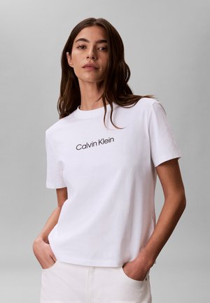 Νεαρή γυναίκα με μακριά καστανά μαλλιά, φορώντας λευκό T-shirt Calvin Klein και λευκό παντελόνι, με τα χέρια στις τσέπες, στέκεται μπροστά σε απλό γκρι φόντο.