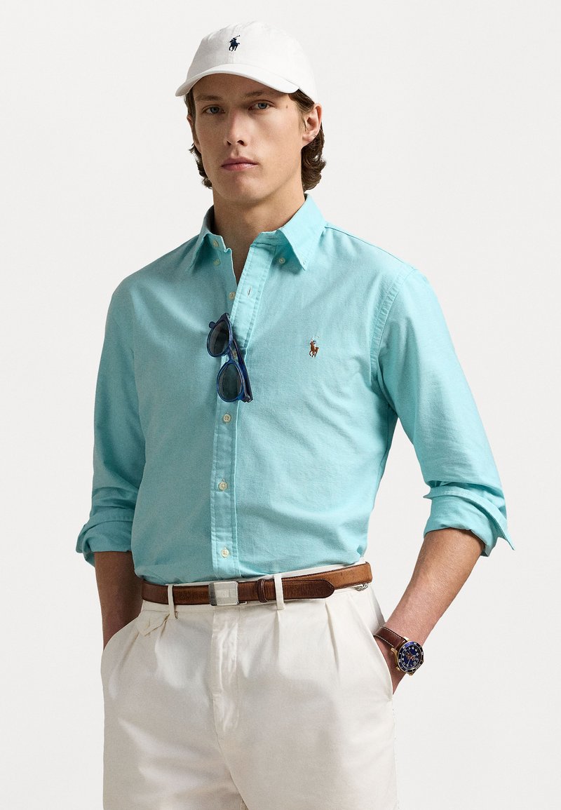 Gai zilas pogkrekls, balta cepure ar logo, brūns josta un gaišas bikses. Aksesuāri ietver saulesbrilles un pulksteni.