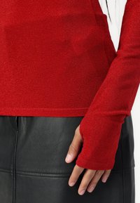 Rotes, langärmliges, figurbetontes Oberteil aus glitzerndem Strickmaterial, mit einem Stehkragen und Daumenlöchern, kombiniert mit einem schwarzen Lederrock.