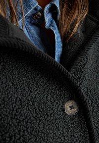 Musta fleece-takki, jossa on teksturoitu pinta, ja siinä on merkittävä hopeinen nappi, johon on kaiverrettu bränditietoja. Denimikuvio näkyy sen alta.