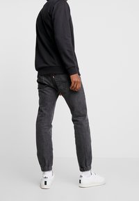 Person wearing black sweatshirt, dark gray jogger jeans with elastic cuffs, white Adidas Stan Smith sneakers, standing on a plain light background.