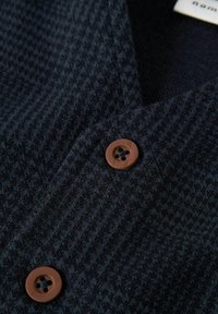 Marineblaues, strukturiertes Gewebe mit einem Hahnentrittmuster, versehen mit zwei braunen Knöpfen. Das Design umfasst einen V-Ausschnitt und eine weiche, gewebte Textur.