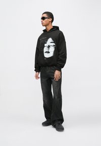 Νεαρός άντρας που φοράει μαύρα γυαλιά ηλίου, μαύρο φούτερ με κουκούλα και λευκή εκτύπωση προσώπου, φαρδιά μαύρα τζιν και μαύρα παπούτσια, στέκεται μπροστά σε ένα απλό φόντο.