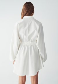 Abito bianco con vita cinchata, caratterizzato da maniche lunghe a palloncino e colletto da camicia. Textura liscia con una vestibilità leggermente oversize.