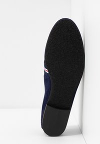 Semelle d'une chaussure en daim bleu foncé avec un dessous en caoutchouc noir texturé et un détail rayé rouge, blanc et bleu sur le côté.