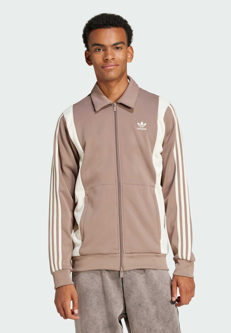 Giacca marrone con zip realizzata in tessuto testurizzato, con pannelli laterali bianchi e tre strisce bianche lungo le maniche. Logo Adidas sul petto.