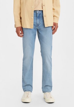 Homme portant un jean droit bleu clair, une chemise beige, une veste beige boutonnée et des baskets écrues, debout devant un fond uni.