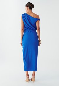 Blauwe asymmetrische jurk met een tekstuurstof, aangesloten bij de taille en uitlopend tot halverwege de kuit. Eenvoudig ontwerp zonder zichtbare patronen of accenten.