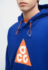 Άνδρας που φοράει μπλε φούτερ με κουκούλα με πορτοκαλί τριγωνικό λογότυπο που περιλαμβάνει λευκά γράμματα «a», «c» και «g», και πορτοκαλί κορδόνια πάνω από πορτοκαλί πουκάμισο.