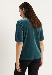 Teal velour kortärmad skjorta med avspänd passform, mjuk textur och rundad halsringning. Synliga sömmar och subtil lyster.