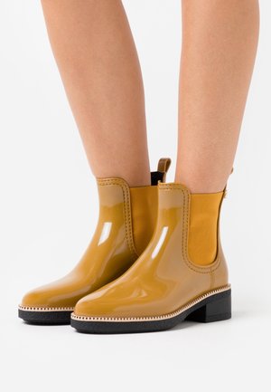 Bottes de pluie chevilles jaune moutarde brillantes avec semelles noires portées sur des jambes nues contre un fond blanc.