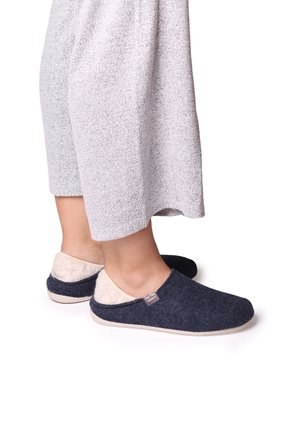 Chaussons en velours marine avec doublure crème, bout rond et semelle légère, associés à un pantalon gris texturé. Design simple à enfiler.