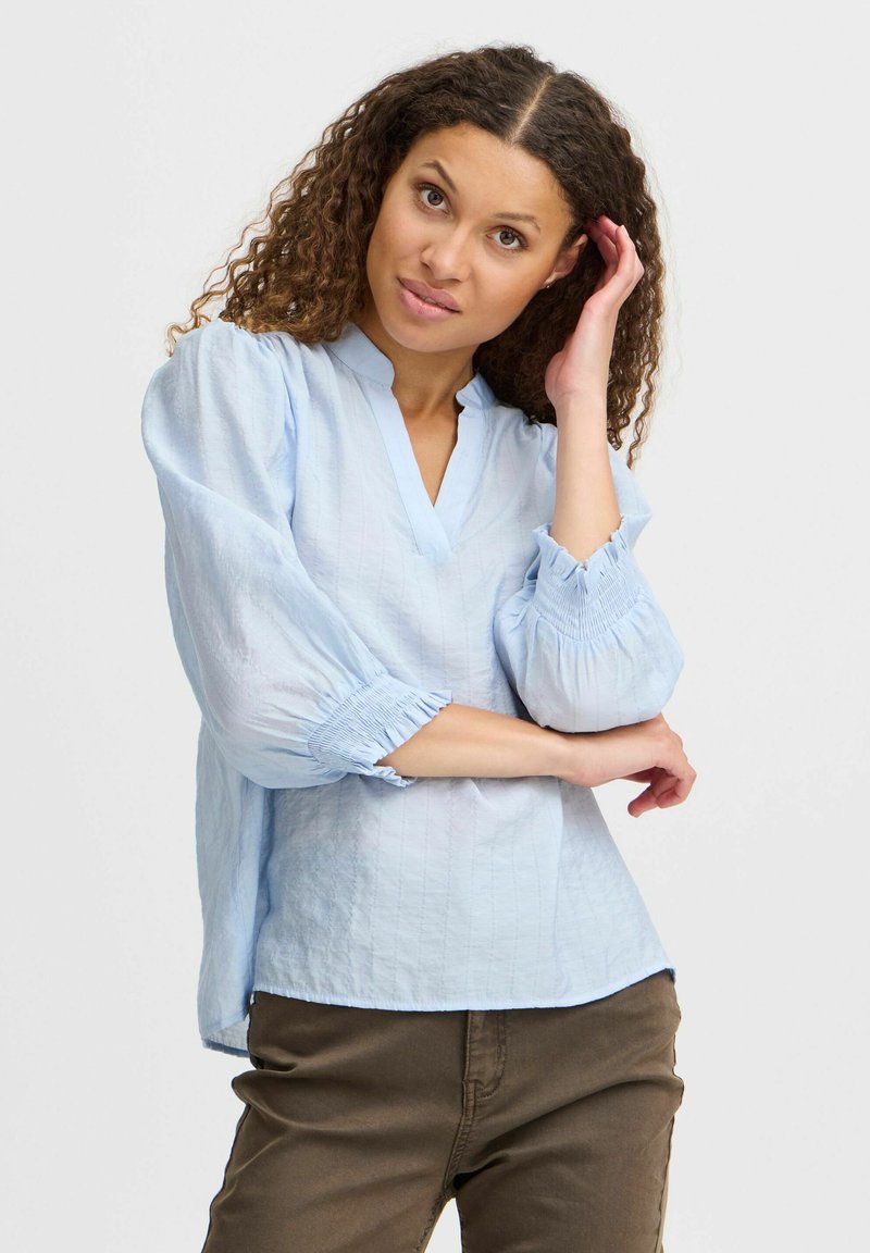 Blusa azzurro chiaro con scollo a V, maniche a sbuffo e polsini arricciati. Realizzata in tessuto testurizzato, presenta una vestibilità comoda e una morbida caduta.