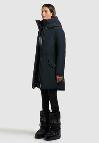 Schwarze isolierte Parka mit hohem Kragen, Reißverschluss vorne und zwei Fronttaschen. Kombiniert mit schwarzen, strukturierten Schneestiefeln und Leggings.