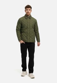Gepolsterte olivgrüne Jacke mit Kragen, Druckknöpfen und Seitentaschen, kombiniert mit schwarzen Hosen und hellen Sneakers.