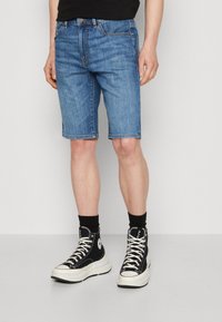 Pantalones cortos de denim en un lavado azul medio, con un corte recto, diseño estándar de cinco bolsillos y dobladillo justo por encima de la rodilla.