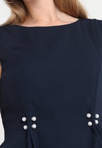 Námořnické modré šaty s lodním výstřihem, zdobené třemi perlovými detaily na každé straně a strukturovaným nařasením v pase.