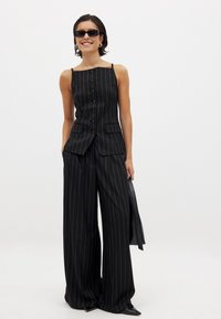 Ženska v črnih prugastih širokih hlačah in ujemajočem brezrokavniku s gumbi, drži črno torbico in nosi sončna očala, se smeji.