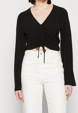 Femme portant un crop top noir à manches longues froncé avec un col en V et un pantalon blanc taille haute sur un fond uni.