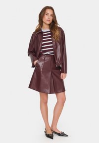 Giacca bomber in pelle bordeaux sopra una maglietta a righe blu e bianche, abbinata a una gonna in pelle bordeaux e a delle scarpette nere a punta.