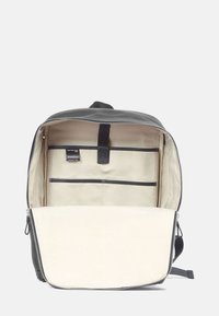 Sac à dos en tissu noir avec une grande ouverture, intérieur beige, plusieurs poches et fermeture à glissière. Design minimaliste avec une étiquette.