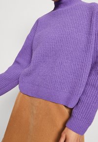 Violetti neulepaita korkealla kaula-aukolla ja tekstuurikuviolla. Päällä ruskea corduroy-hame, jossa pystysuorat ribbitukset.